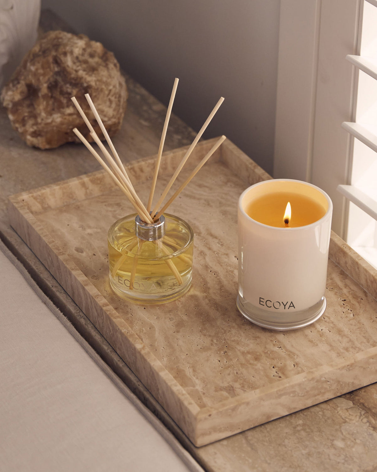 Lit Ecoya Madison candle beside a reed diffuser on a travertine tray