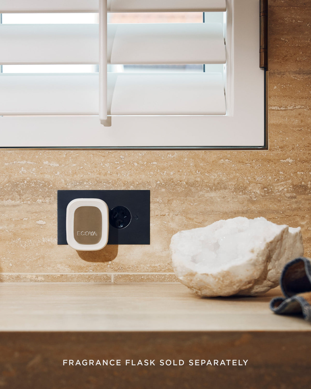 Ecoya Plug-In Diffuser in a black socket in a tiled bathroom below white shutters, beside a crystal