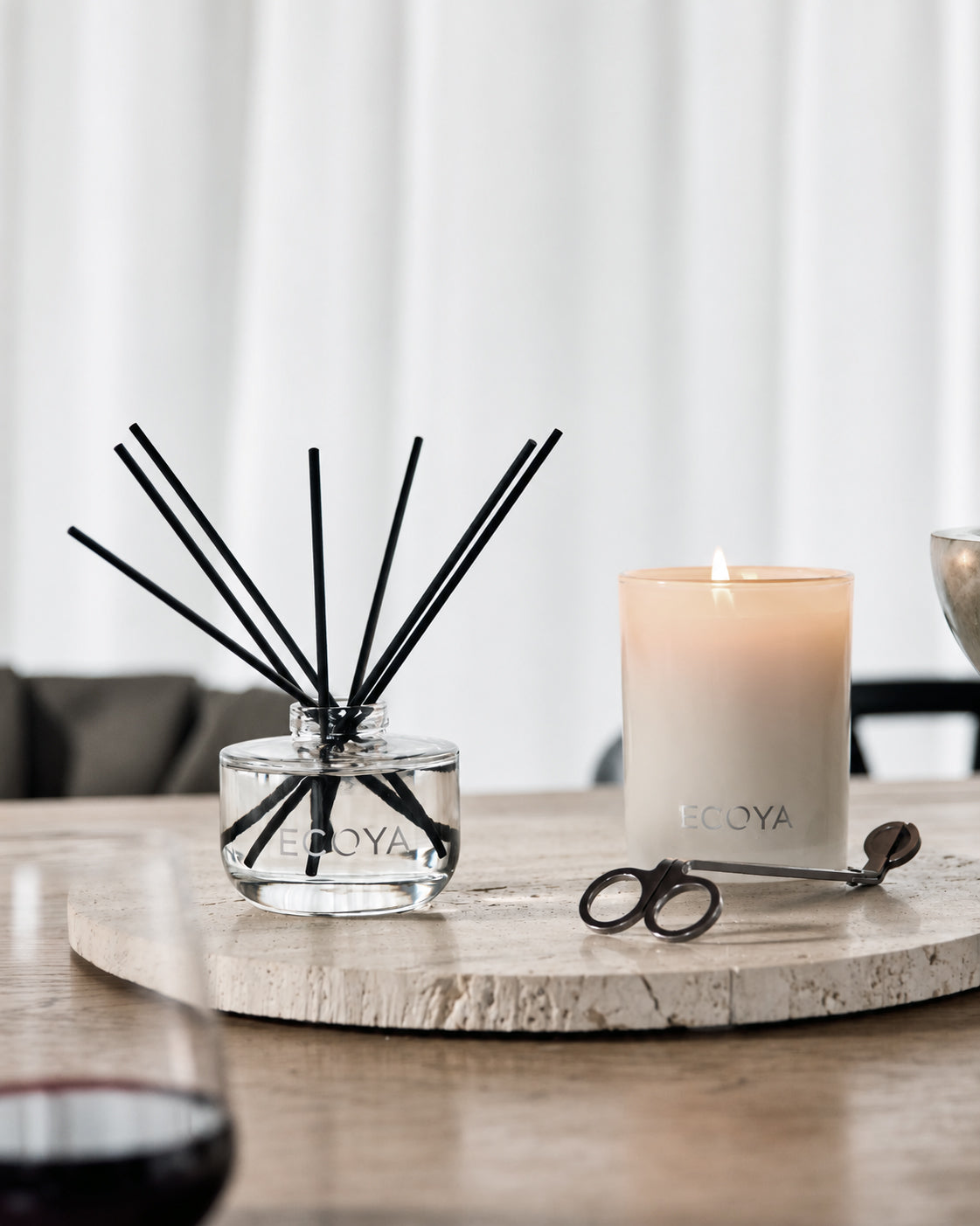 Ecoya reed diffuser and lit Ecoya candle with a wick trimmer on a round travertine tray, on a timber table beside a glass of red wine
