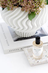 Ecoya room spray bottle on a terrazzo tray in front of a large white fluted vase filled with pink flowers, styled on top of a neutral coffee table book.