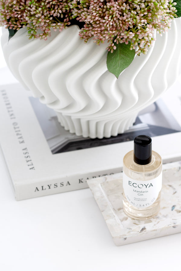 Ecoya room spray bottle on a terrazzo tray in front of a large white fluted vase filled with pink flowers, styled on top of a neutral coffee table book.
