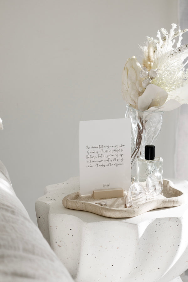 A set of affirmation cards displayed on a neutral bedside table with a wooden stand, dried floral arrangement, and perfume bottle styled in a soft, minimalist bedroom.