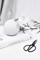 Black bonsai pruning shears resting on a white surface beside The Kinfolk Table book and a round white vase holding a single blossom stem.