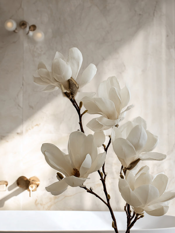 Close-up of ultra-realistic white magnolia stems with wide cupped petals in soft natural light, styled in front of a marble wall and brass bathroom tapware for a dreamy, refined look.