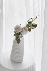 A single, realistic faux cabbage rose stem in a soft blush-ivory hue, elegantly displayed in a modern, white ribbed vase. The simple arrangement sits on a white side table against a sheer white curtain, creating a light, airy, and romantic minimalist scene.