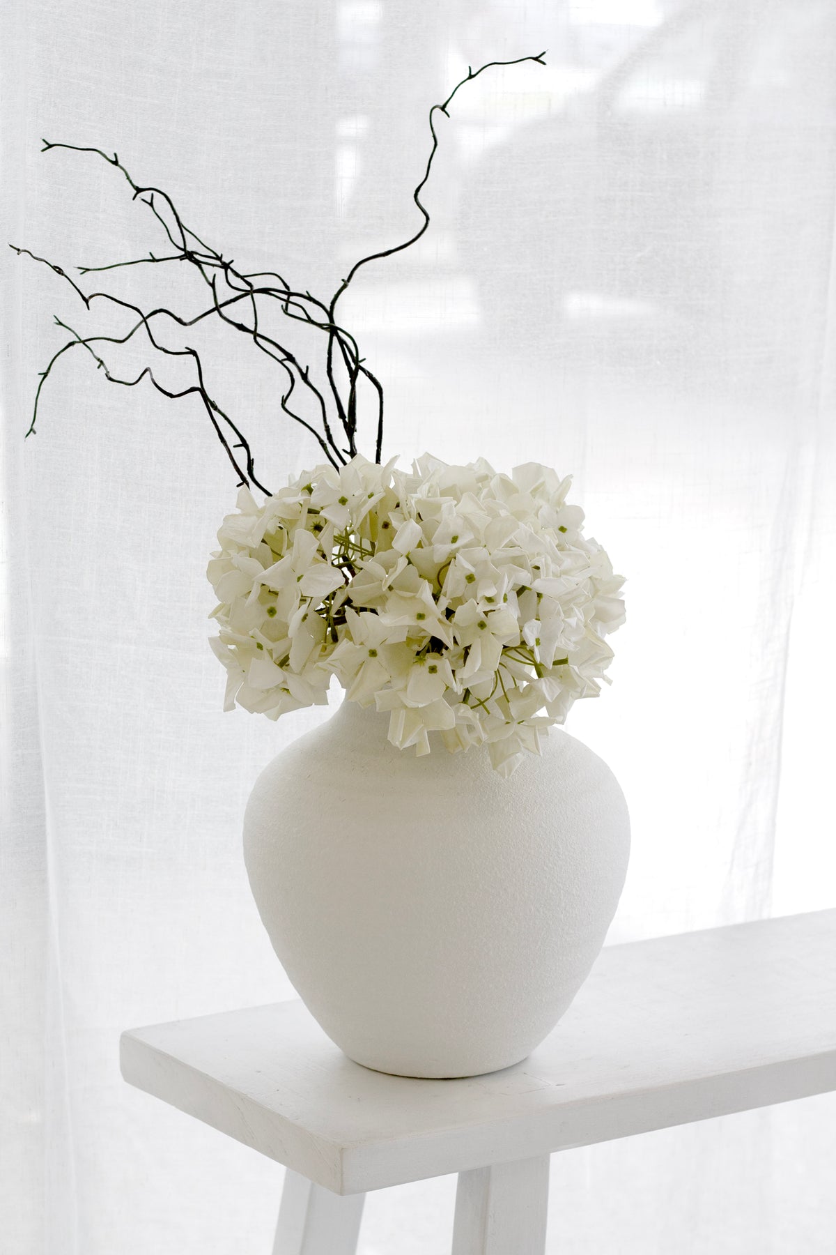 Cream faux country hydrangea with dark twigs in a textured white vase on a white stool