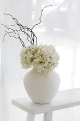 A striking and minimalist floral arrangement featuring a realistic white faux country hydrangea, paired with dark, sculptural twigs for dramatic contrast. The arrangement is set in a matte white, textured vase on a simple white stool against a bright, sheer curtain, creating an artistic, high-contrast, and serene aesthetic.