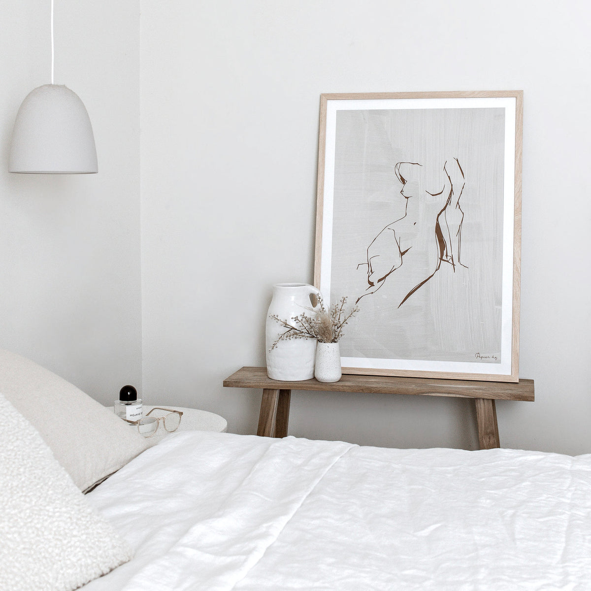 Figure print in a natural frame on a timber bench above a bed with white linen