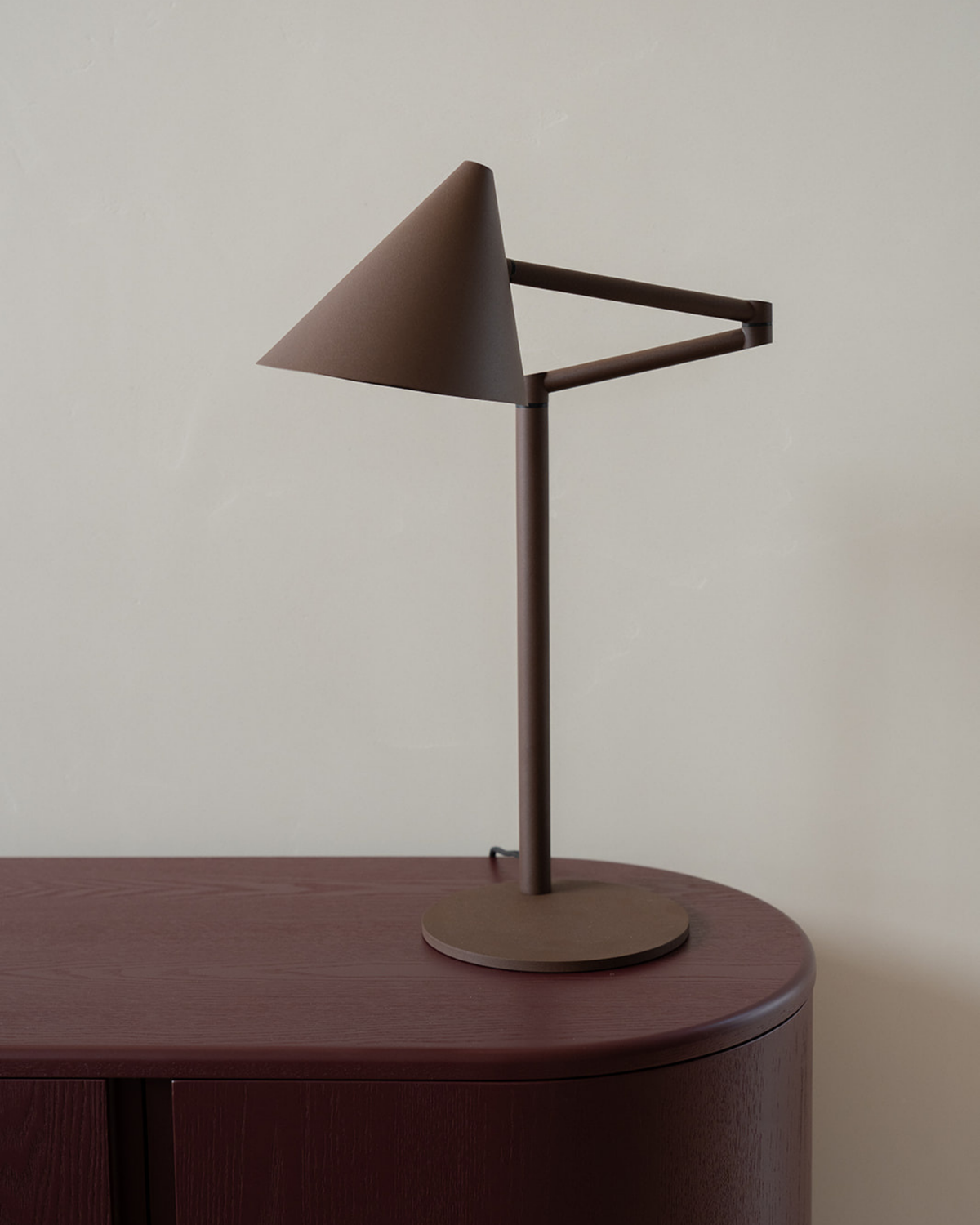 Coffee Brown lamp on the curved end of an oxblood sideboard, its conical shade and folded arm side on