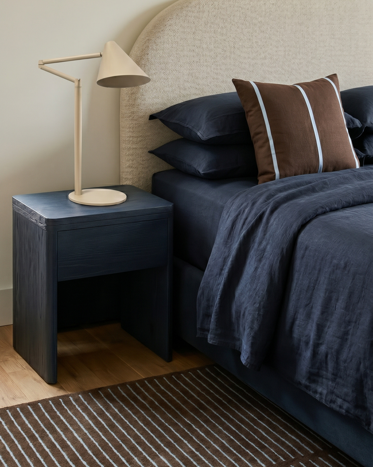 Oat lamp on a navy timber bedside table next to a bed with navy linen and a brown striped cushion