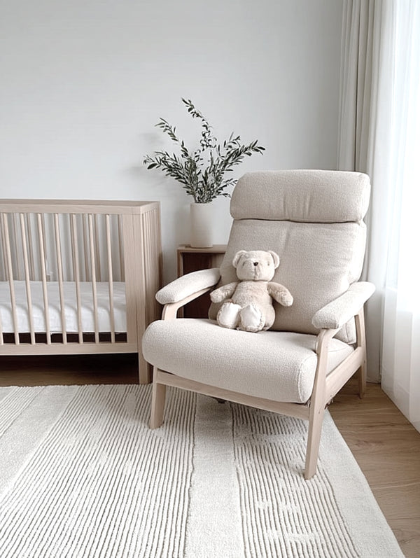 Modern nursery featuring a cream armchair with a soft plush bear toy, a timber cot, potted greenery in a ceramic vase, and a textured white rug on light timber floors.