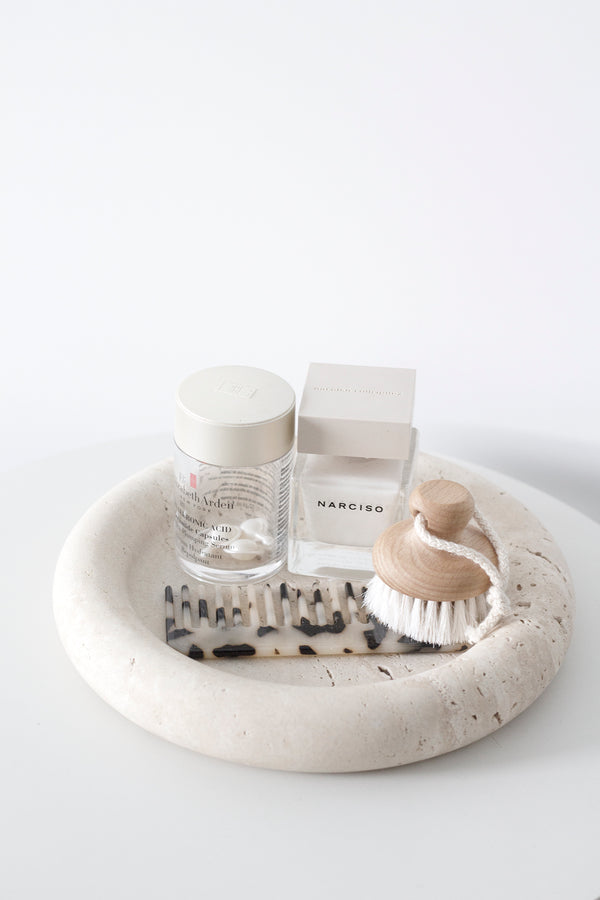 Natural goat hair face brush with wooden handle styled on a travertine tray beside Narciso perfume, skincare serum, and a tortoiseshell comb in a minimal bathroom vignette.