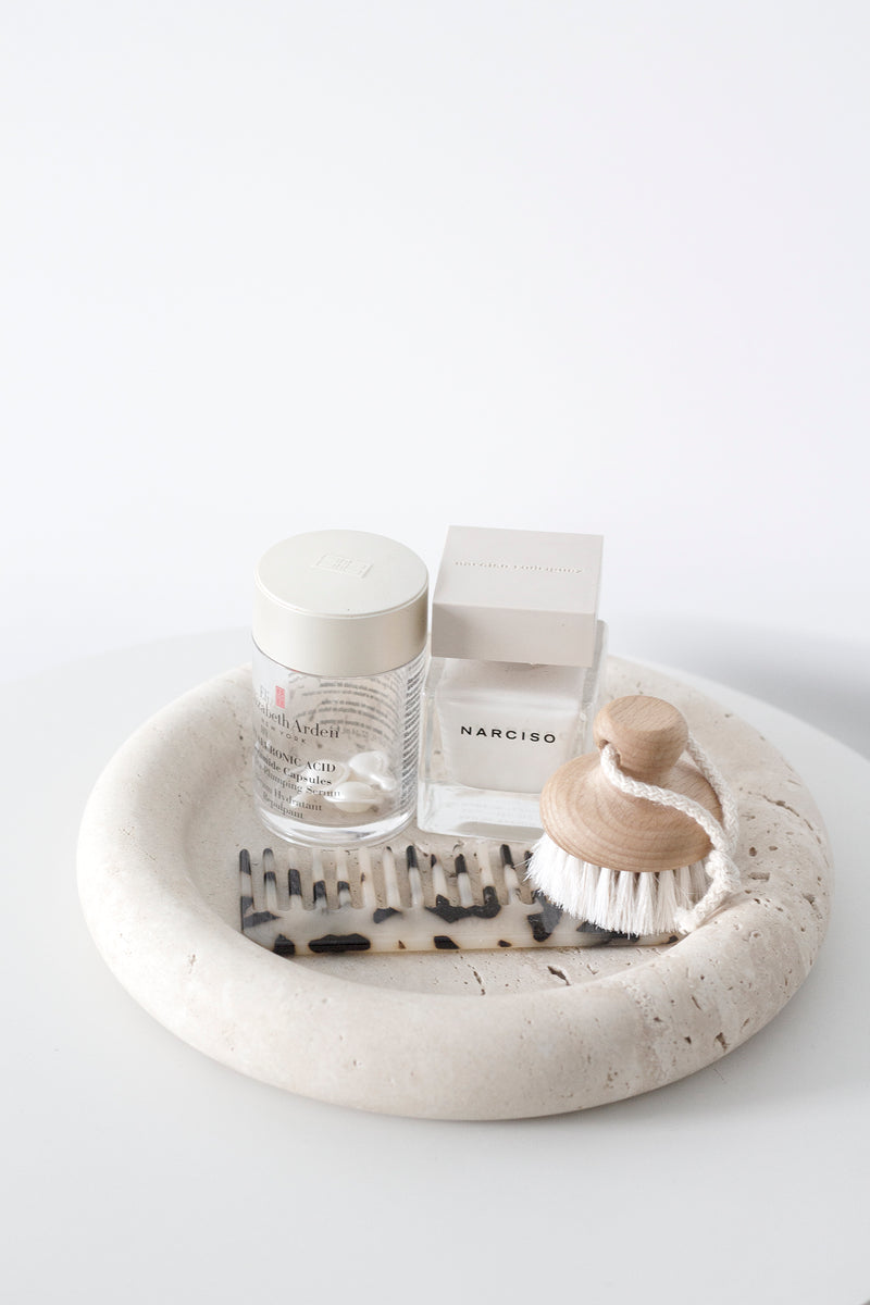 Natural goat hair face brush with wooden handle styled on a travertine tray beside Narciso perfume, skincare serum, and a tortoiseshell comb in a minimal bathroom vignette.