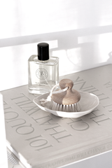 Florence Brushware goat hair face brush with a wooden handle on a small white dish, styled on a coffee table book beside a clear perfume bottle in soft sunlight