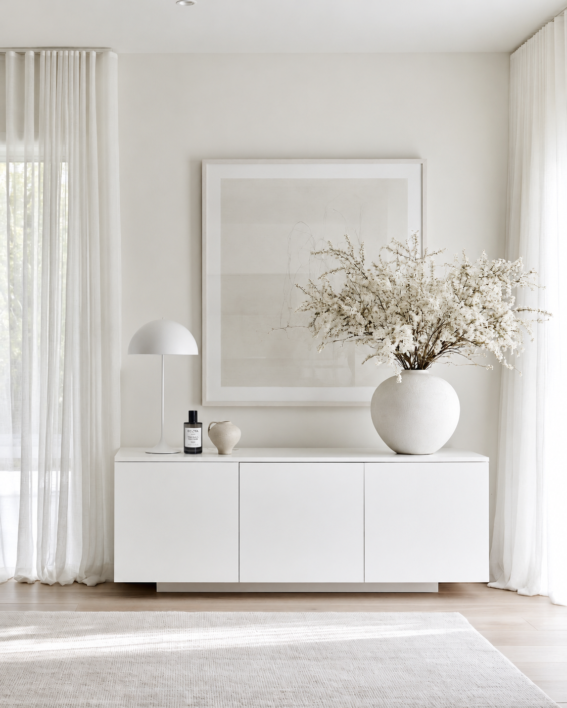 Ecoya Celebration room spray on a white sideboard beside a lamp and a vase of blossom