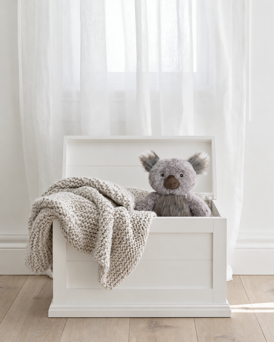 Kenny Koala mini soft toy by Lily & George peeking out of a white wooden toy box beside a knit blanket