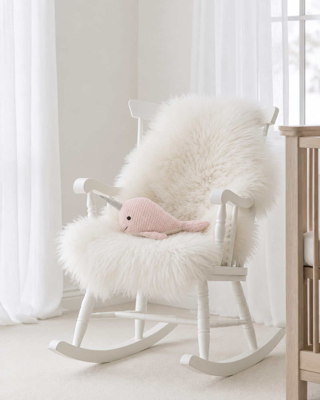 Nellie Narwhal knitted rattle by Lily & George lying on a sheepskin on a white nursery rocking chair