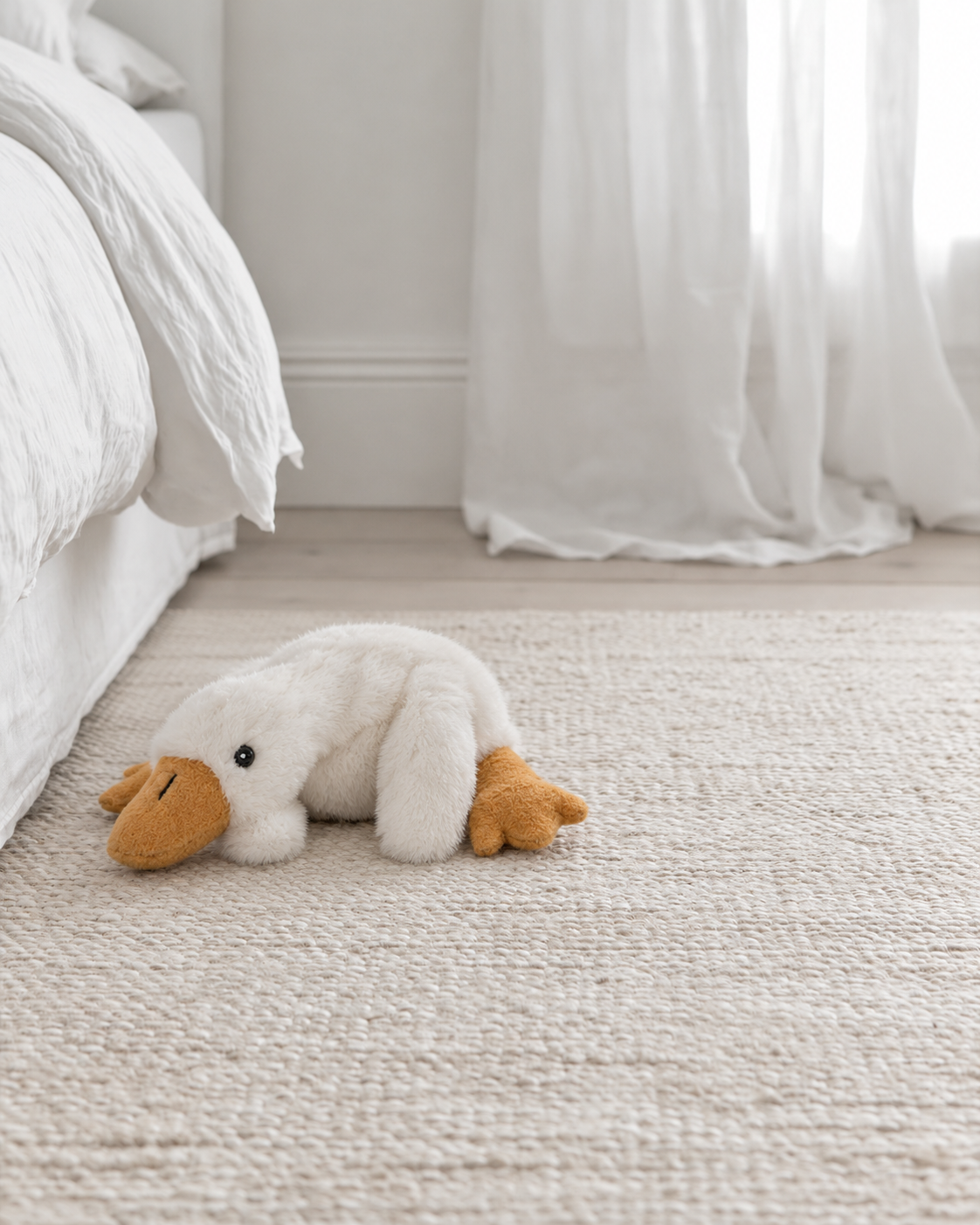 Millie the Goose soft toy by Lily & George lying on a textured wool rug beside a white bed