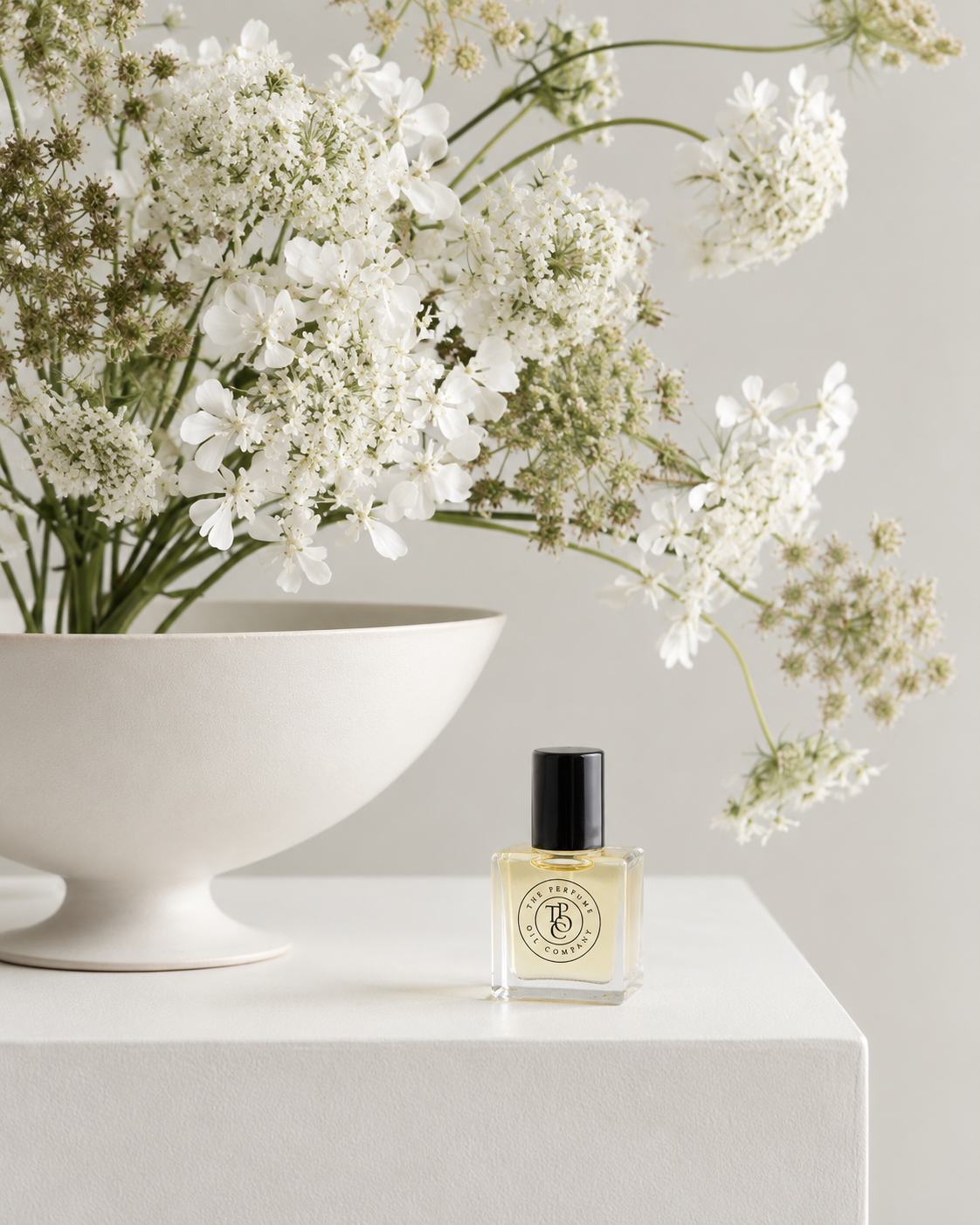 DAMSEL Perfume Oil on a white plinth beside the bowl of white flowers