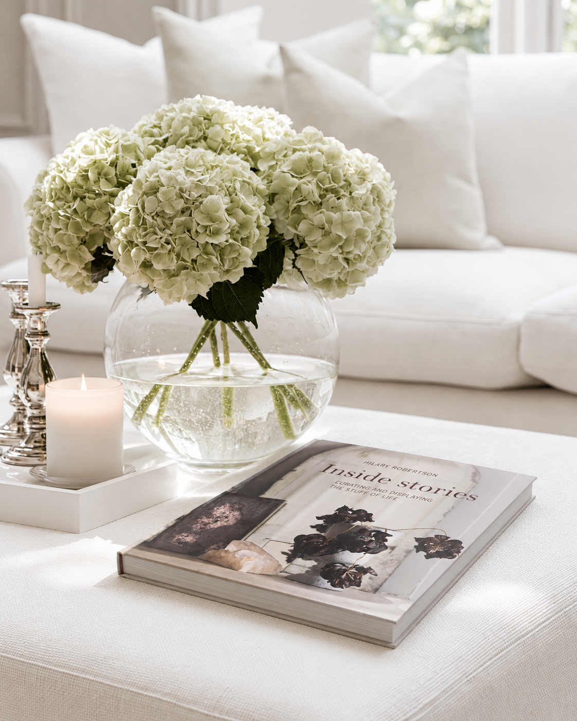 Inside Stories by Hilary Robertson on a white ottoman beside a vase of green hydrangeas
