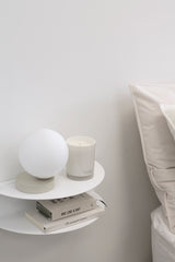 White FOLD wall-mounted bedside table styled with a lamp, candle, and books beside a neutral linen bed.
