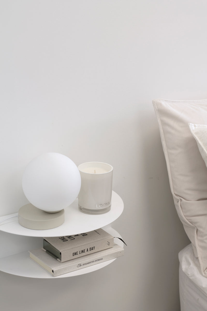 White FOLD wall-mounted bedside table styled with a lamp, candle, and books beside a neutral linen bed.
