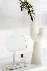 Off-white sculptural Franki vase with cactus-like shape holding green berry stems, styled on a white console beside a small white mirror and perfume bottle in front of sheer curtains.