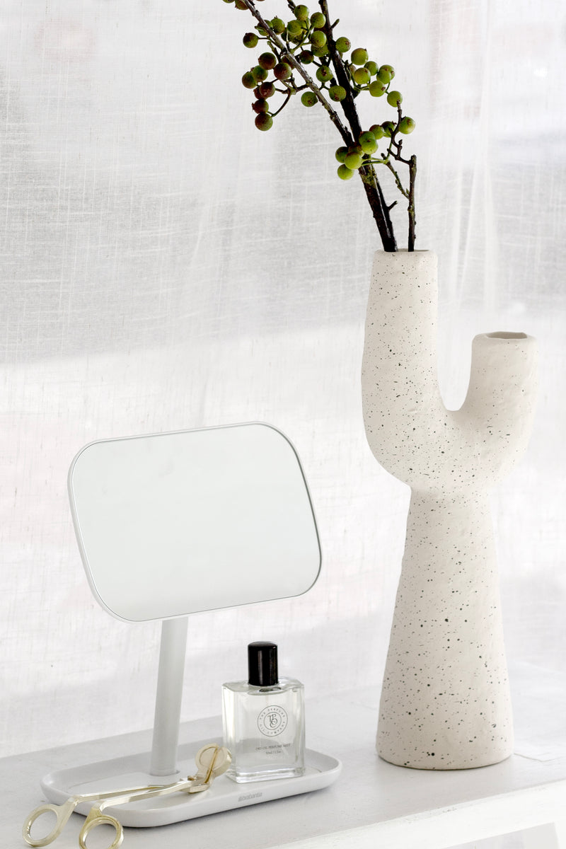 Off-white sculptural Franki vase with cactus-like shape holding green berry stems, styled on a white console beside a small white mirror and perfume bottle in front of sheer curtains.
