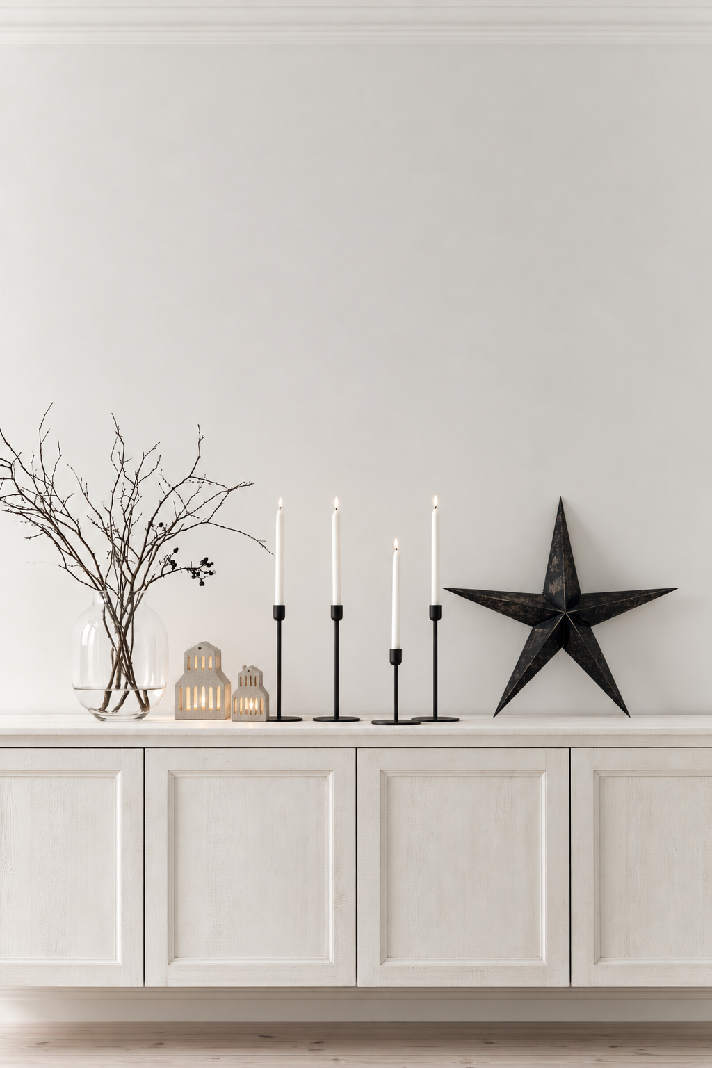 Two gable roof house tealight holders glowing on a white sideboard beside black candlesticks and a star
