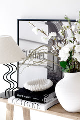 Long faux willow branch styled through a white vase with white blossoms on a console with a sculptural lamp, framed art and stacked fashion books