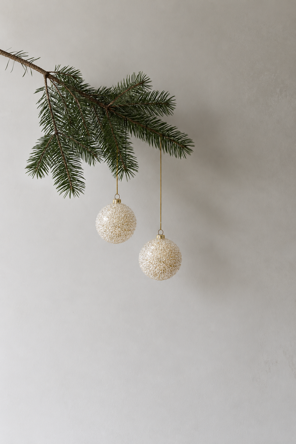 Two gold beaded baubles hanging from a pine branch against a soft grey wall