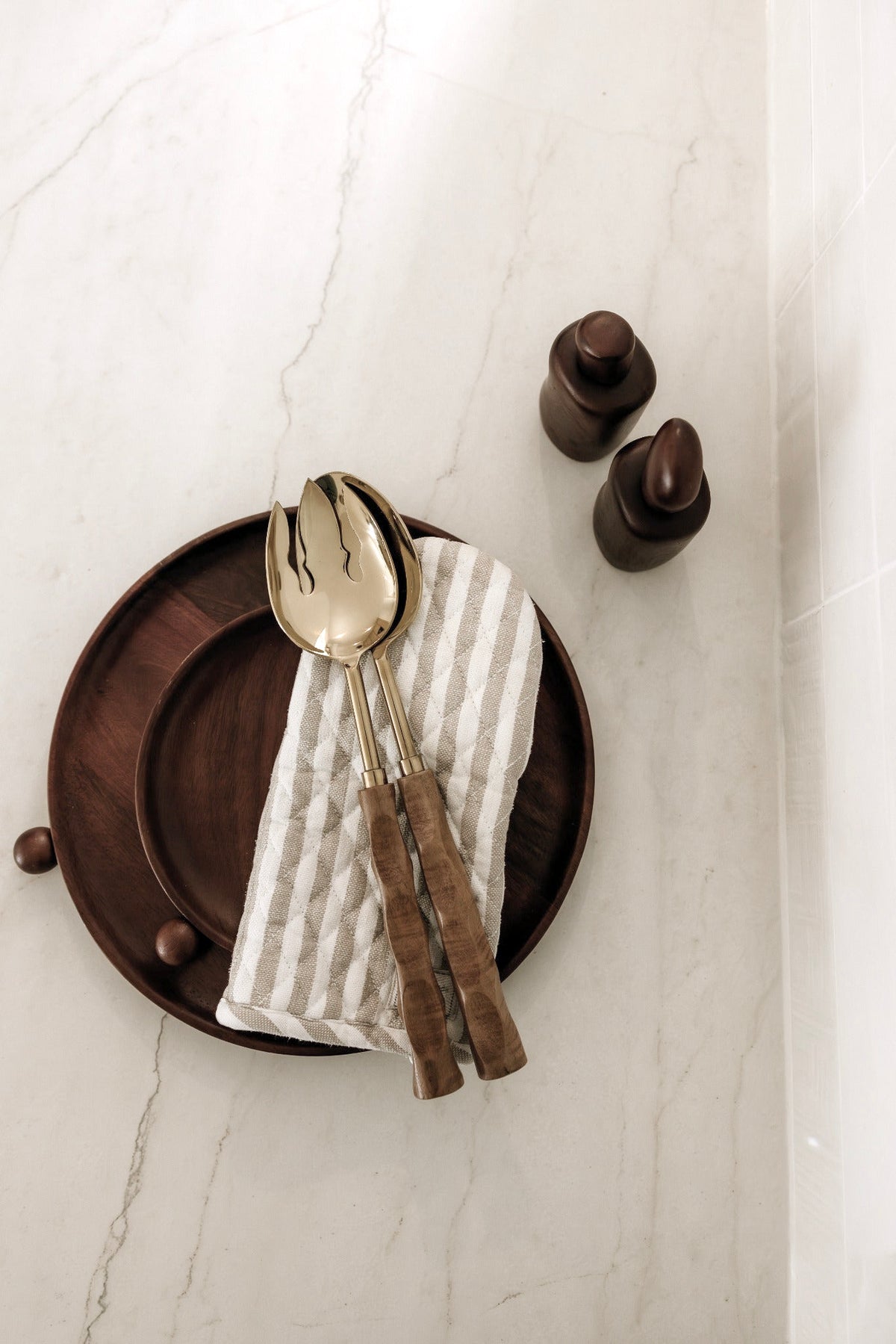 Gold and wood salad servers on a linen napkin and dark plate beside wooden shakers