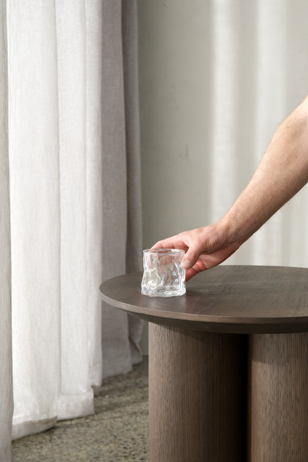 Hand setting a rippled clear glass tumbler on a round timber side table beside linen curtains