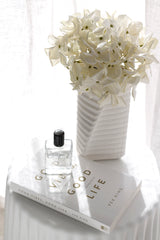 Cream grandiflora hydrangea faux blooms arranged in a tall white ribbed vase, styled on a white book with a perfume bottle in soft sunlight