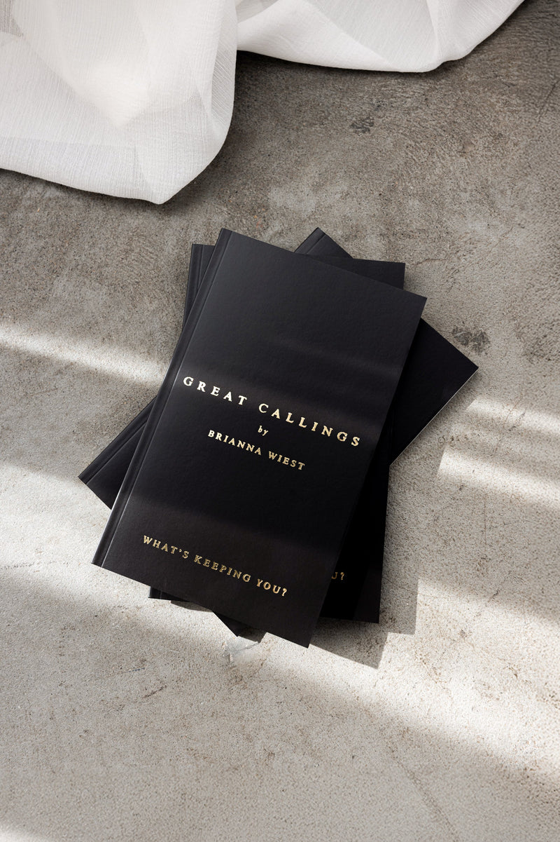 Flatlay of Brianna Wiest’s black and gold book Great Callings on a concrete surface beside soft white fabric in natural light.