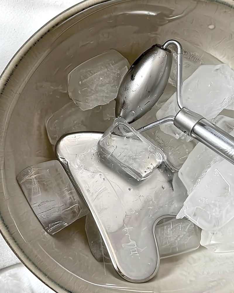 Close-up of a stainless steel gua sha tool and facial roller resting on ice cubes in a ceramic bowl, perfect for cooling and de-puffing skin.