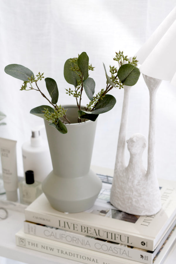 Hamburg Vase in sage ceramic with classic tapered silhouette styled with faux greenery on books beside sculptural lamp.
