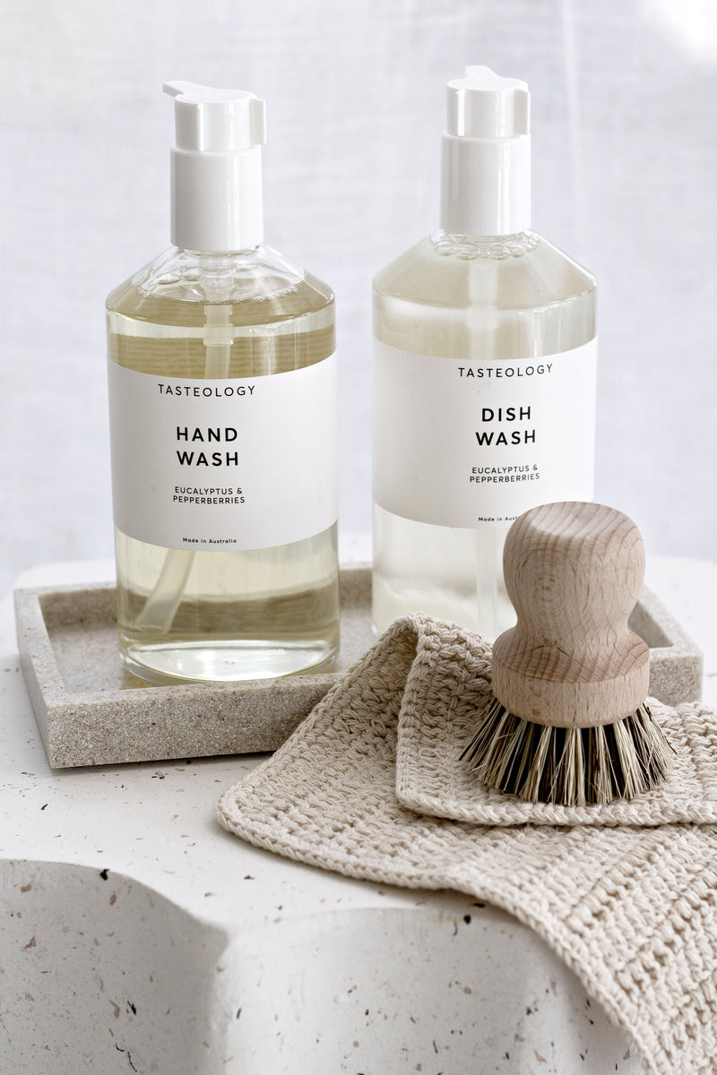 Handmade cotton face cloth in unbleached natural tone, styled with Tasteology hand wash, dish wash, and wooden scrubbing brush on stone surface.