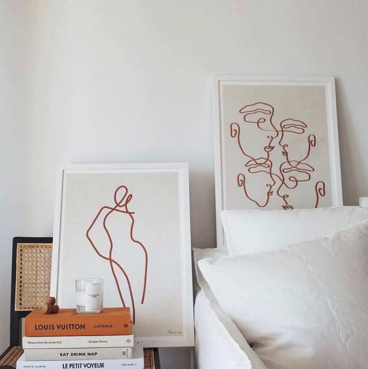 Homebodii Linen print in rust framed beside a line art faces print on a stack of books