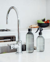 Ecoya Juniper Berry and Mint fragranced dish liquid and surface spray in matte sage green bottles on a white kitchen bench beside a chrome gooseneck tap with fresh produce in background