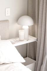 Ecoya Celebration candle in White Musk and Warm Vanilla in a ribbed white glass jar, styled on a bedside table beside a white dome lamp and neutral curtains