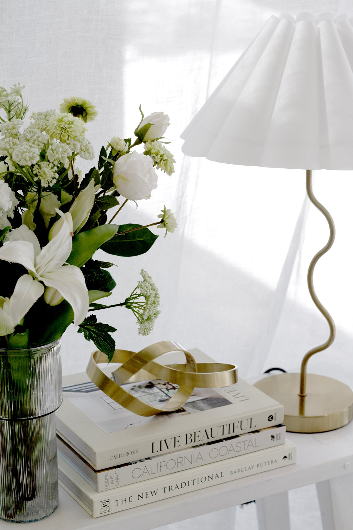 Brushed gold Infinity Loop sculpture on coffee table books beside a lamp and white flowers