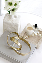 Brushed gold Infinity Loop Sculpture styled on a grey Dior Catwalk book with a white vase of flowers, marble tray, candle, and perfume bottle, creating a chic modern vignette.