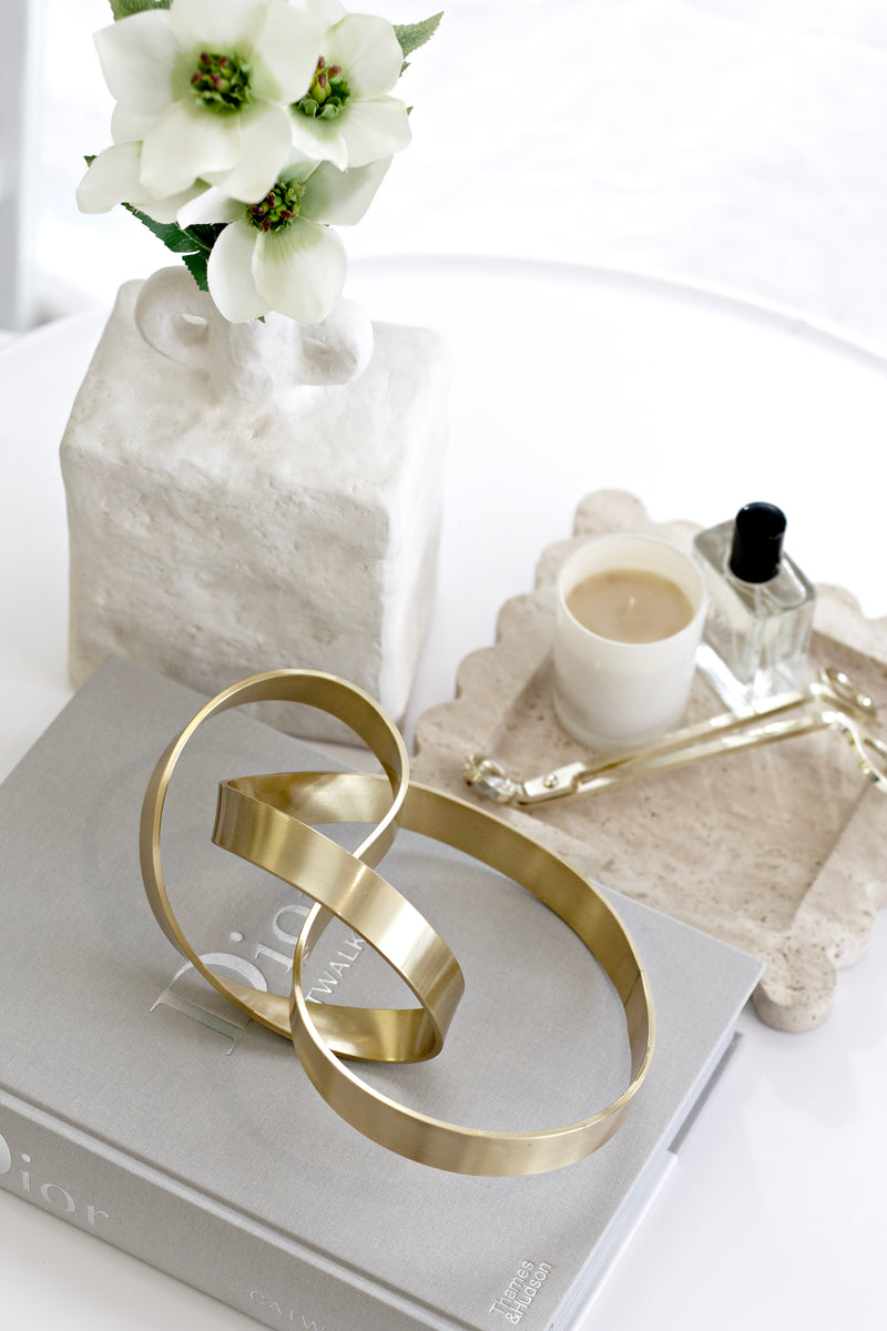 Brushed gold Infinity Loop Sculpture styled on a grey Dior Catwalk book with a white vase of flowers, marble tray, candle, and perfume bottle, creating a chic modern vignette.