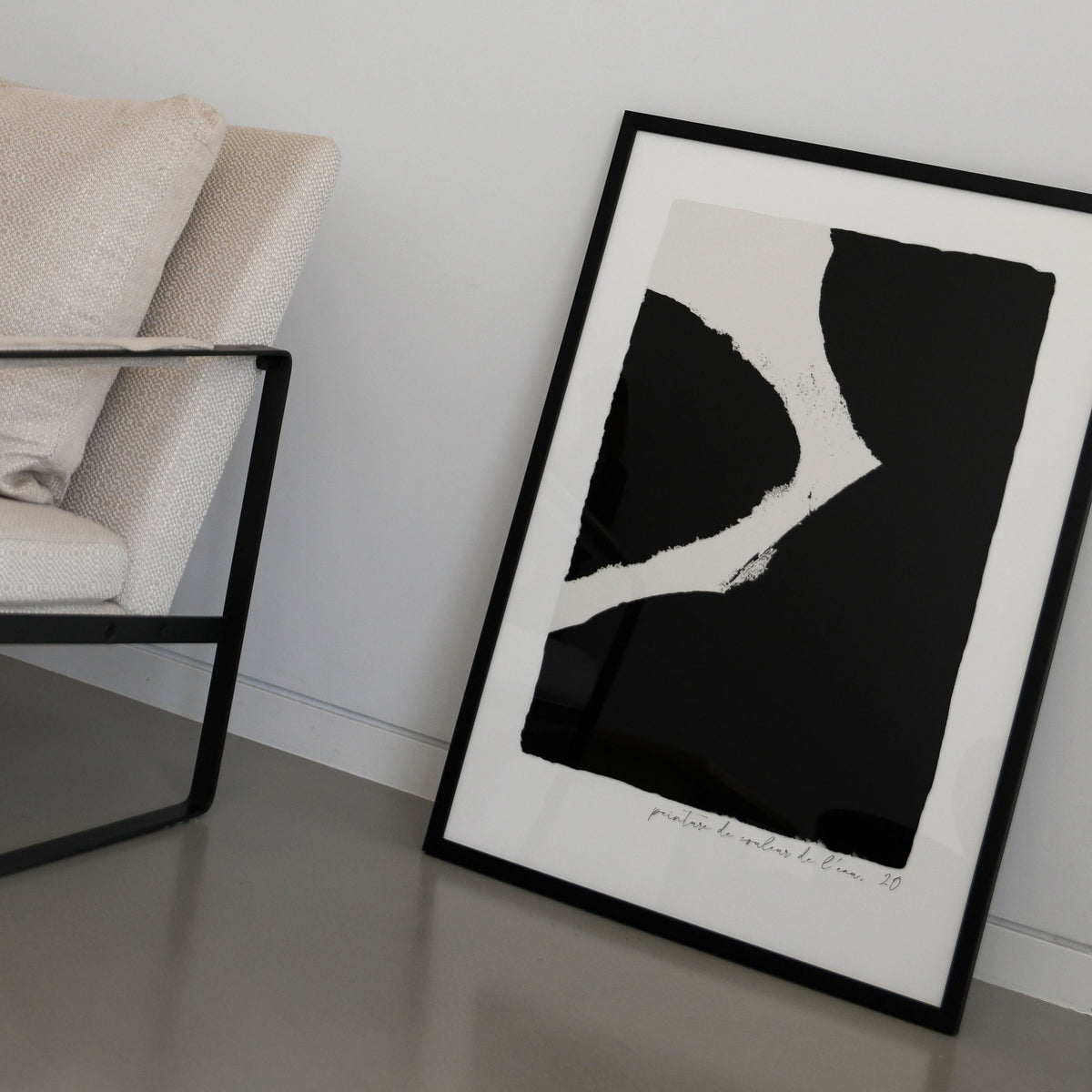 Ink print in black in a black frame leaning on the floor beside a cream armchair
