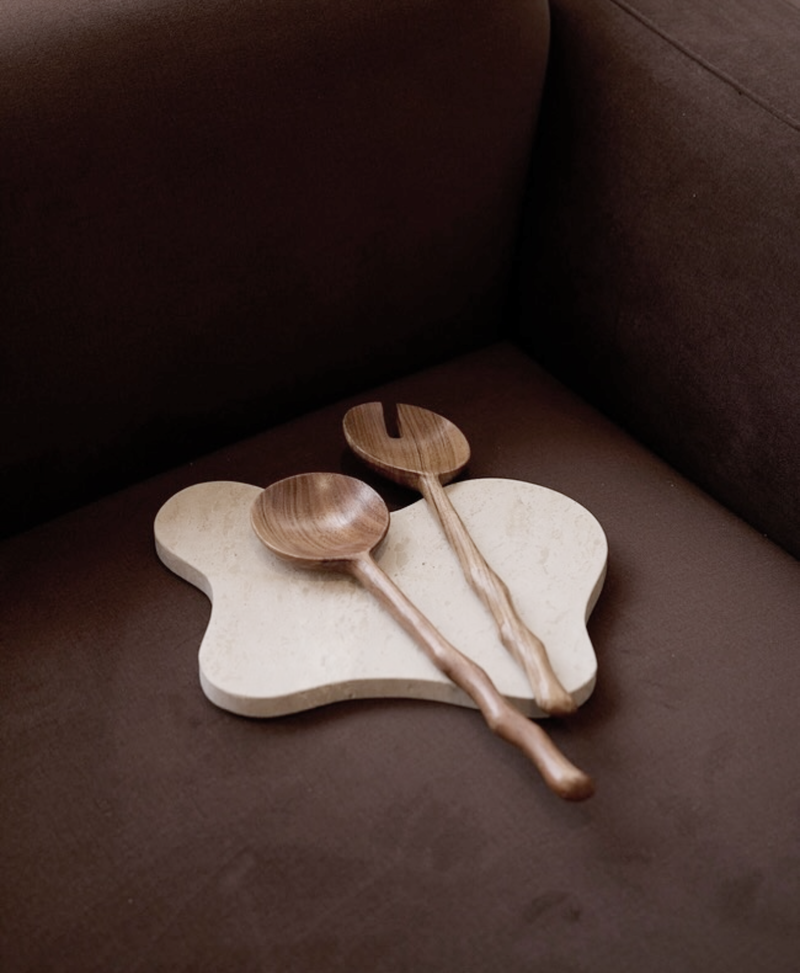 Isobel Travertine Tray with sculptural organic shape styled with wooden serving utensils on a dark sofa.