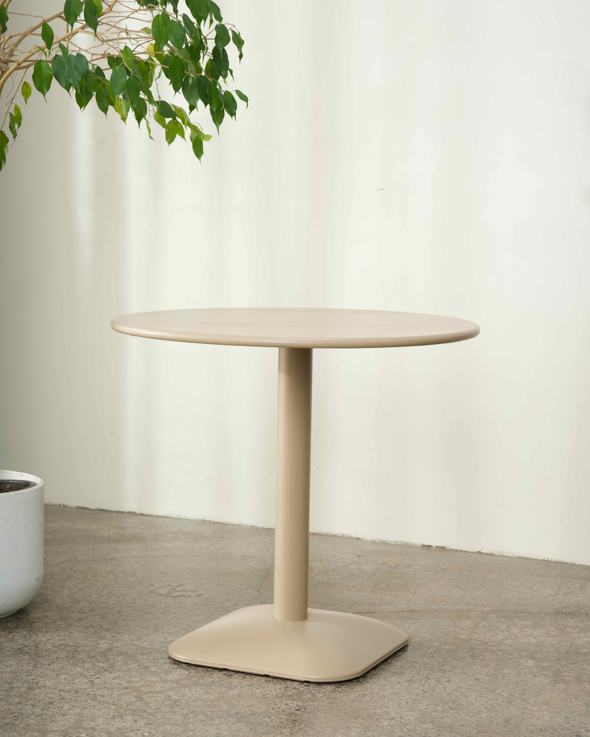 Jasper Table by Ned Collections in Nude, an 80cm round steel top on a single column and squared foot, beside a fig tree
