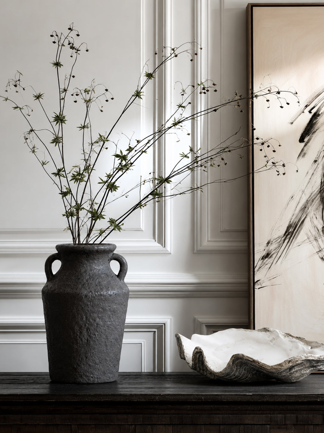 Black Kaia stone vase of blossom branches beside a white resin clam shell dish on a dark console