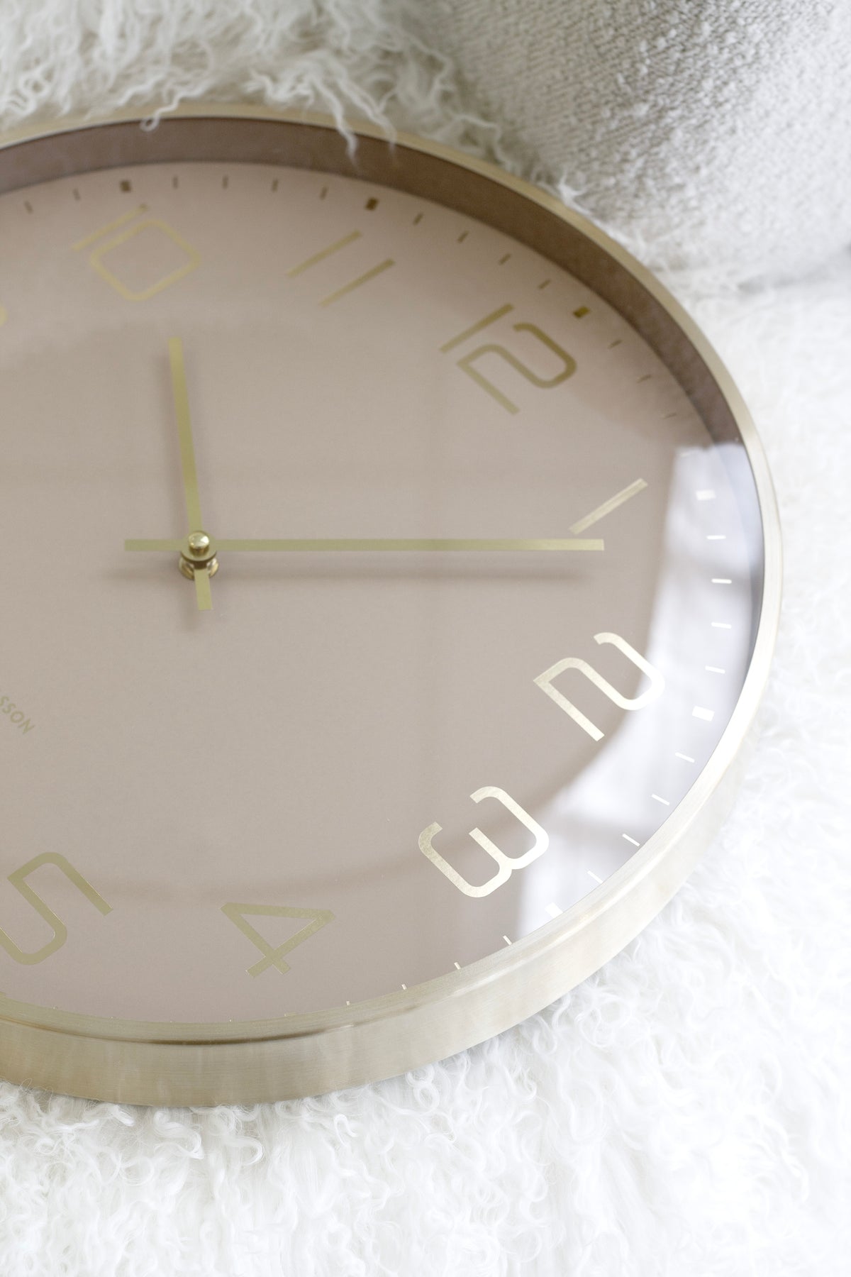 Soft brown wall clock with gold hands and modern numerals resting on white textured fabric