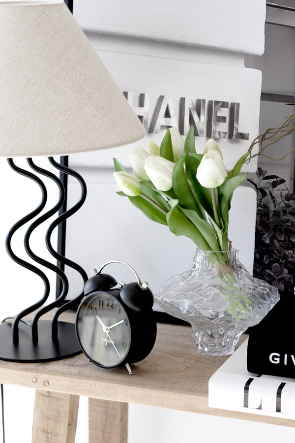 Karlsson Iconic Alarm Clock in black styled on bedside table with lamp, flowers, and books.